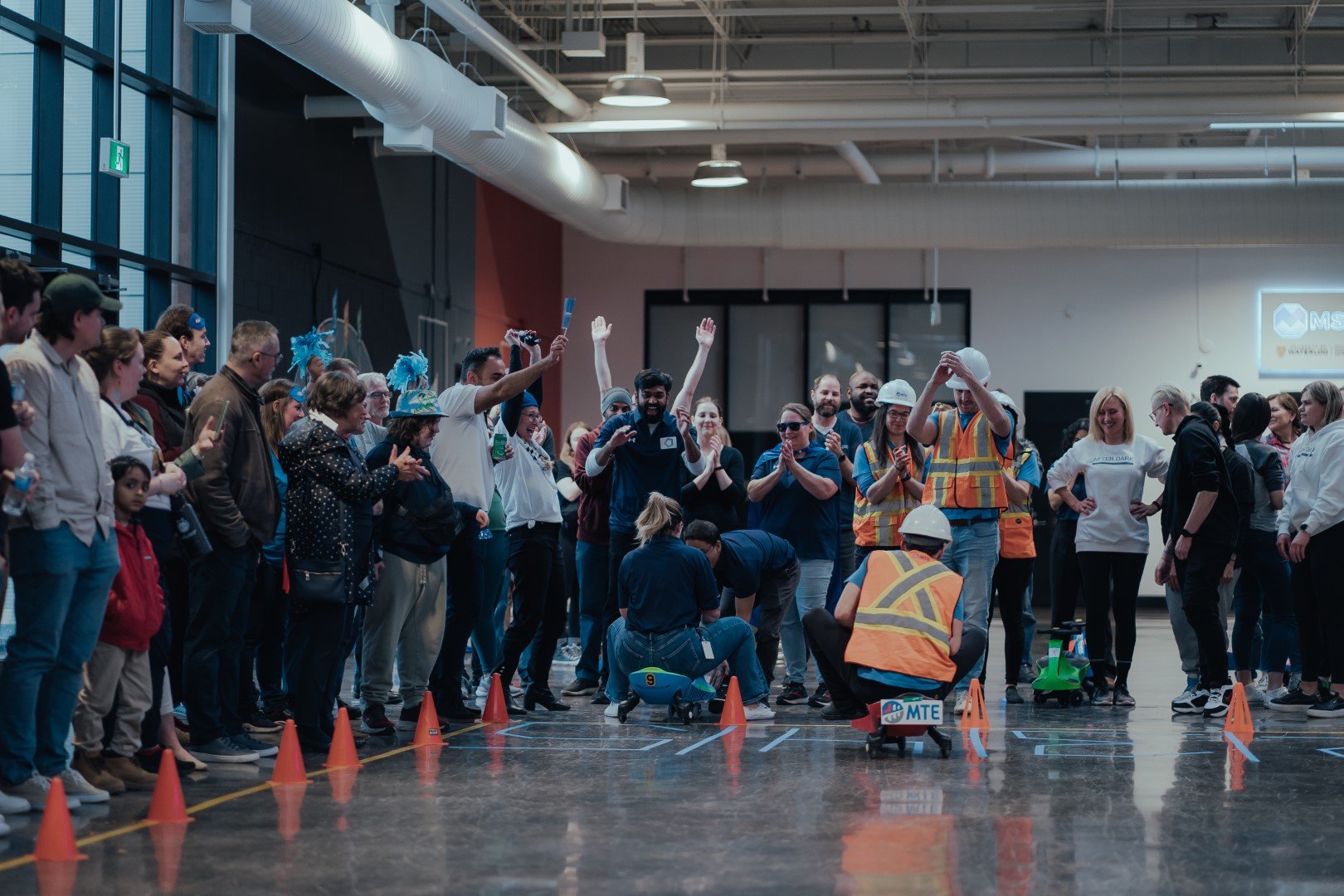 A large group of people watches and cheers as participants in safety vests, highlighting MTE community involvement, ride small wheeled carts inside a spacious indoor venue surrounded by orange cones and industrial decor.
