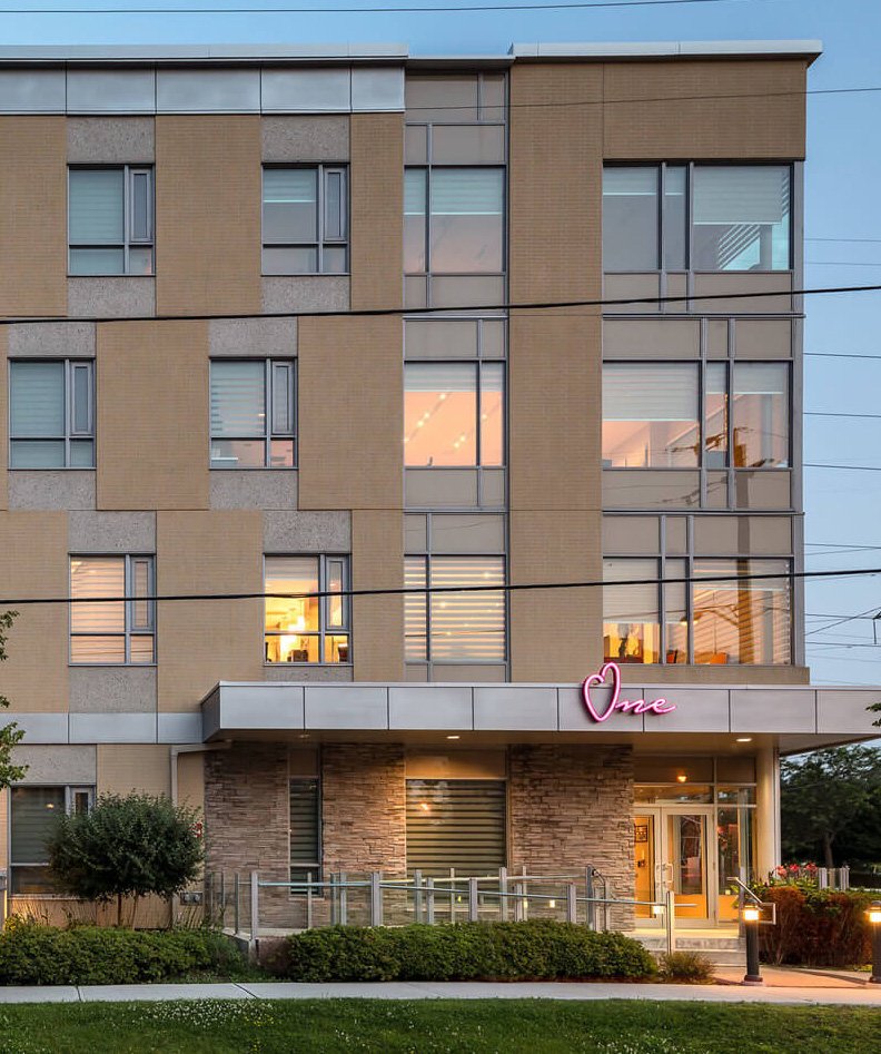 A modern, four-story building with large windows, tan and gray exterior, and a neon pink sign that says One above the entrance—supporting the delivery of long-term care homes in Ontario. The ground floor features stone accents and landscaped greenery.
