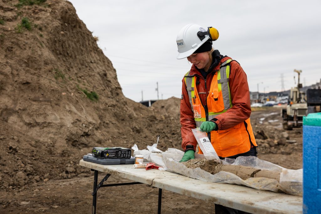 A construction worker in safety gear examines soil samples on a table at an outdoor work site, supporting the MTE Kitchener Central Transit Hub project, with mounds of dirt and equipment visible in the background.