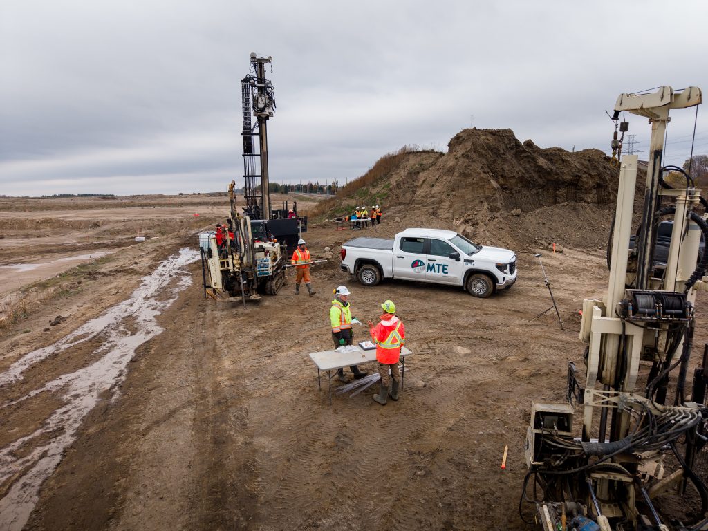 Several workers in safety gear operate drilling equipment at a construction site with dirt mounds and machinery. An MTE-branded pickup truck, supporting the Kitchener Central Transit Hub, is parked nearby under a cloudy sky.